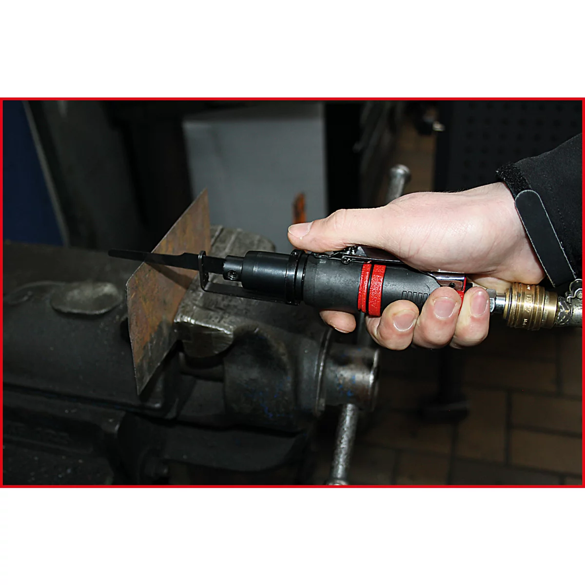 Hand hält eine Stichsäge, schneidet Metallplatte. Die Säge ist schwarz mit roten Akzenten. Die Metallplatte wird in einem Schraubstock gehalten.